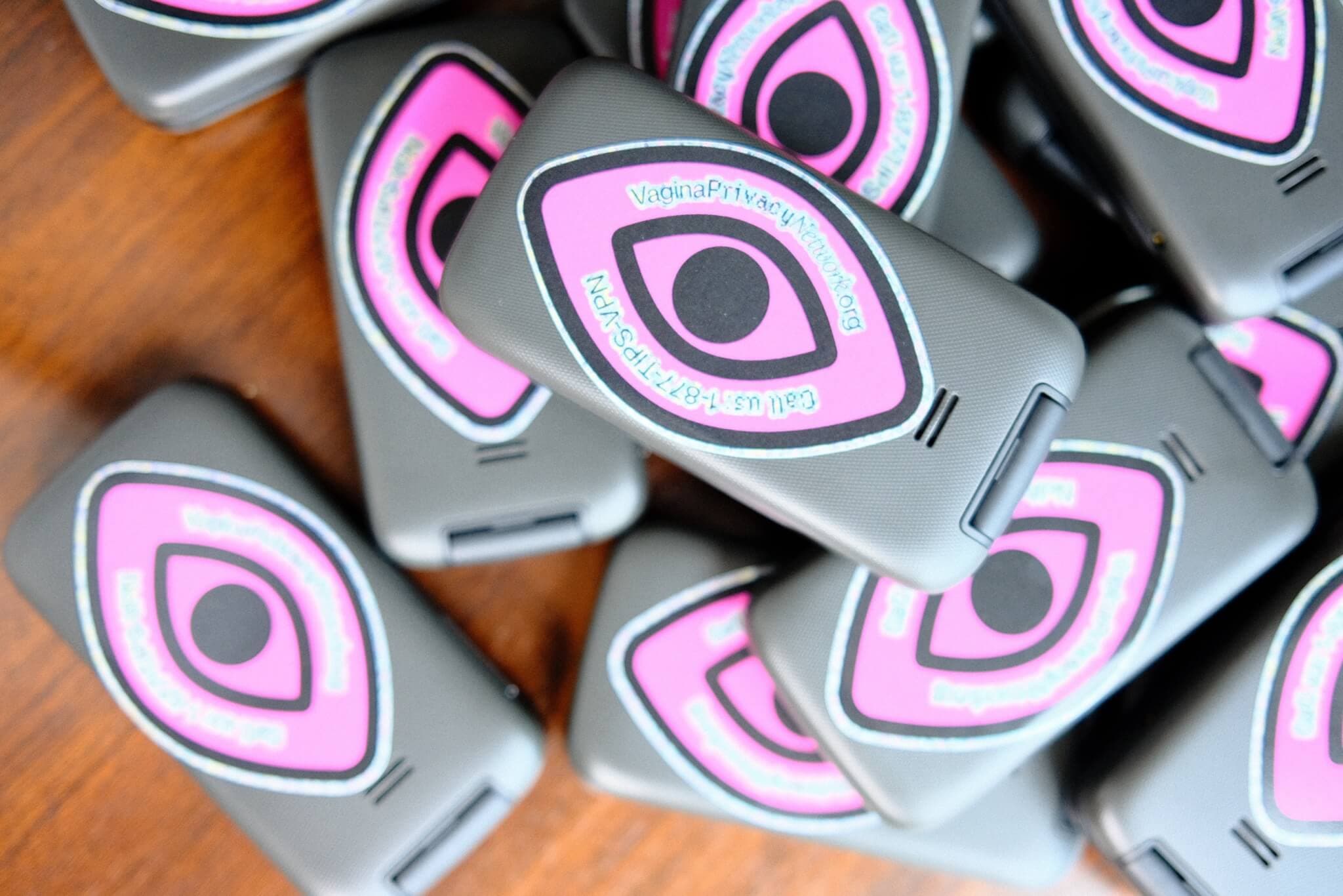 A stack of phones with a pink and black sticker reading "VaginaPrivacyNetwork.org. Call us 10977-TIPS-VPN"